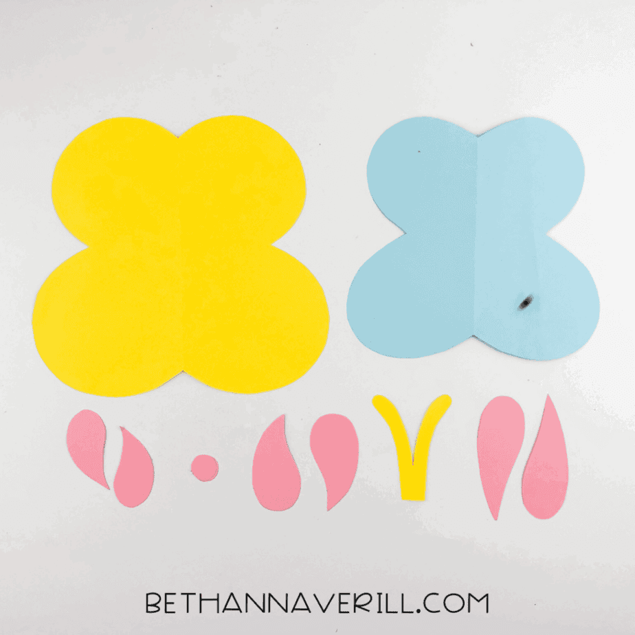 Cut cardstock pieces arranged on a white background, including yellow and blue butterfly wing shapes, pink wing details, and small yellow antenna pieces.
