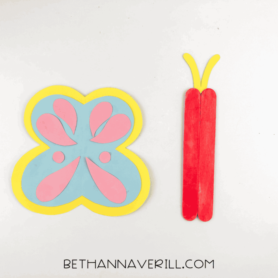 Assembled butterfly wings placed next to the pink-painted craft stick body with yellow antennae.