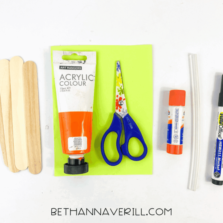 Overhead view of craft supplies on a white background including three natural wooden craft sticks, a tube of orange acrylic paint, child-safe scissors, a glue stick, two clear glue sticks, and a black marker, arranged neatly beside a piece of bright green cardstock.