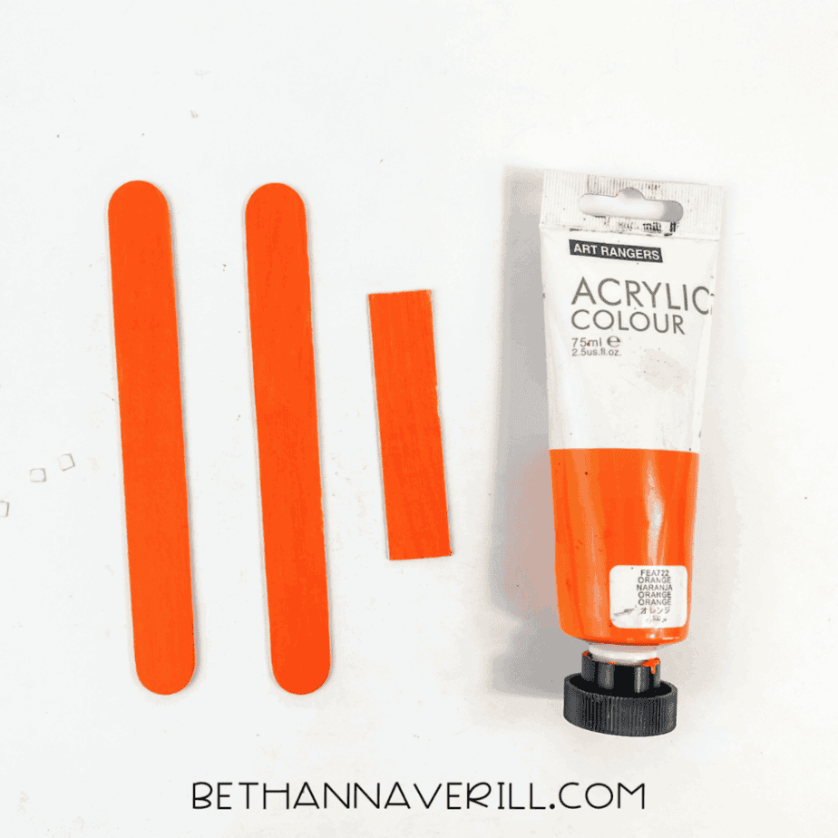 Two full-length wooden craft sticks and one shorter cut piece painted orange, placed horizontally on a white background next to a tube of orange acrylic paint.