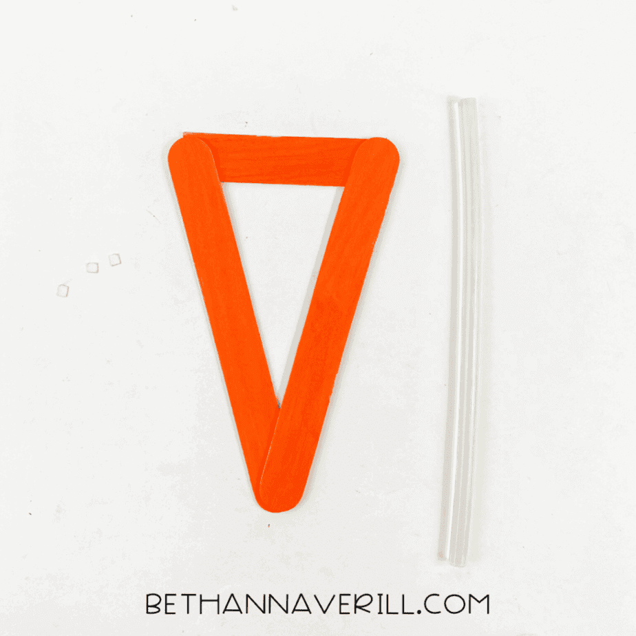 Three orange-painted craft sticks glued together to form a triangular shape resembling a carrot outline, placed on a white background beside two clear glue sticks.