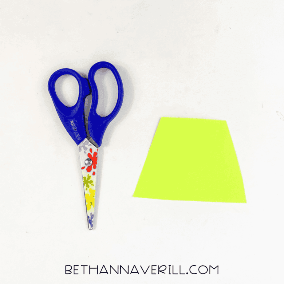 A pair of child scissors next to a trapezoid-shaped piece of bright green cardstock on a white background.