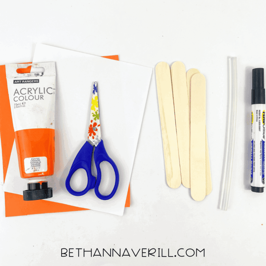 Flat lay of orange acrylic paint, child-safe scissors, white and orange cardstock, four plain wooden craft sticks, two hot glue sticks, and a black marker on a white background.