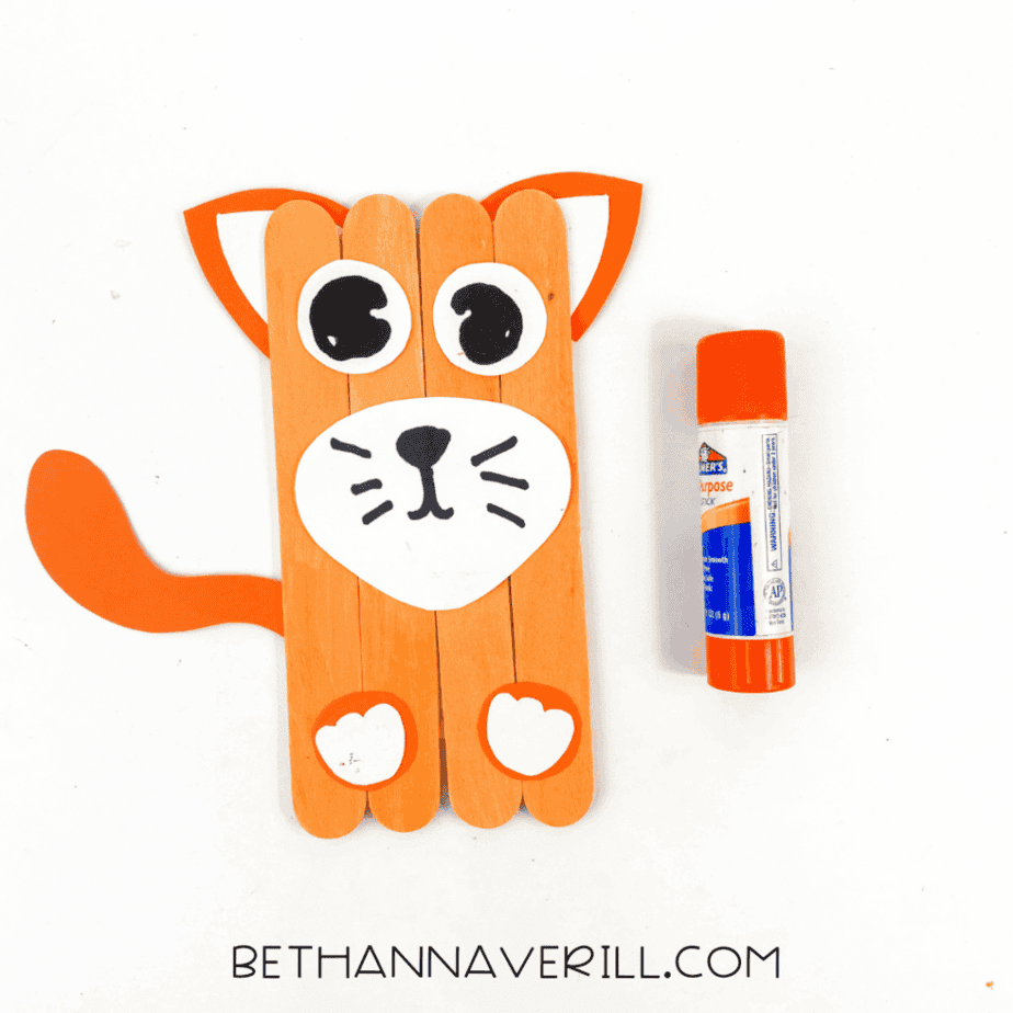 Cat craft with orange paper tail attached to the left side and white paw shapes added at the bottom, glue stick placed to the right.