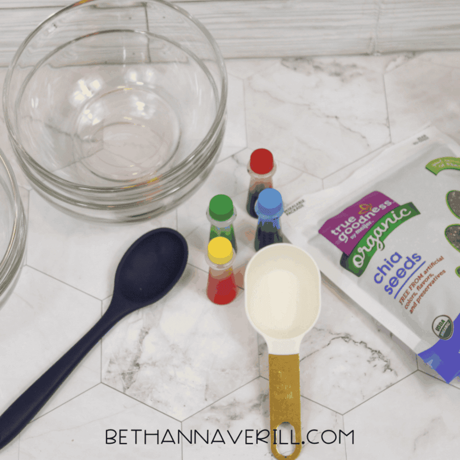 Overhead view of sensory bin supplies on a white surface including glass bowls, chia seeds in a measuring cup, liquid food coloring bottles, a large spoon, and a bag of organic chia seeds.