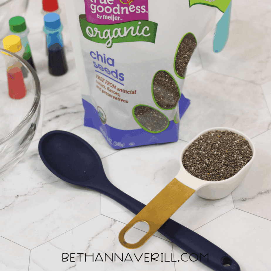 Measuring cup filled with dry chia seeds resting on a large spoon, placed next to a bag of organic chia seeds on a white surface.