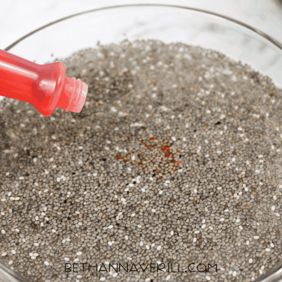 Close-up of yellow liquid food coloring being dropped onto hydrated chia seeds in a glass bowl.