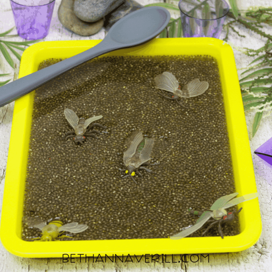 Yellow plastic sensory bin filled with hydrated chia seeds and plastic insect toys, with a large spoon resting across the top.