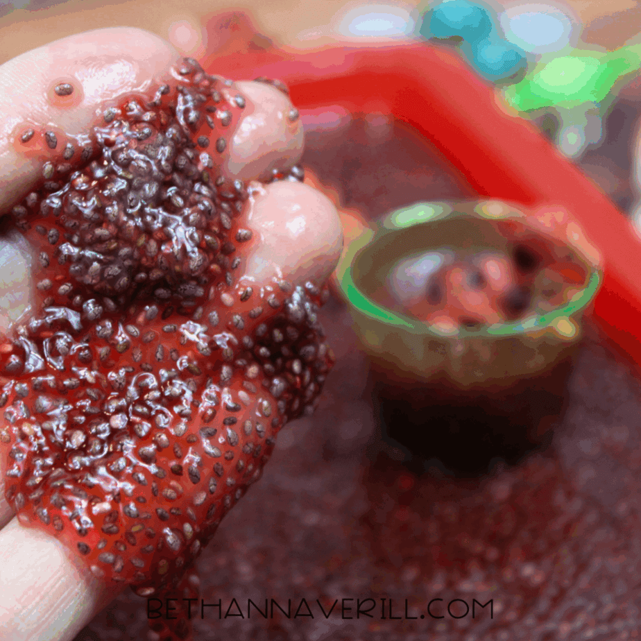 Close-up of hydrated chia seeds dripping from a small clear cup over a sensory tray.