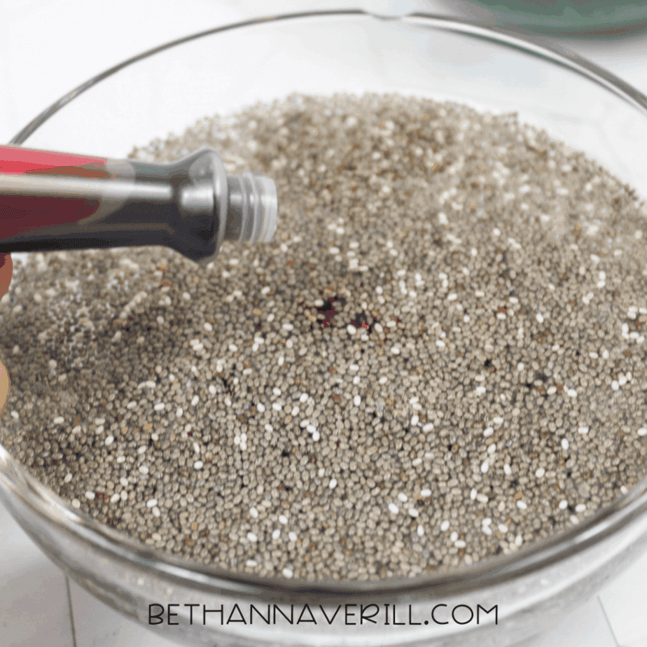 Close-up of red food coloring being poured onto dry chia seeds in a glass bowl.