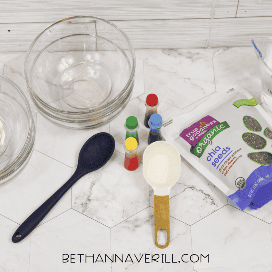 Supplies arranged on a countertop, including glass bowls, food coloring bottles, measuring cups, a spoon, and a bag of chia seeds.
