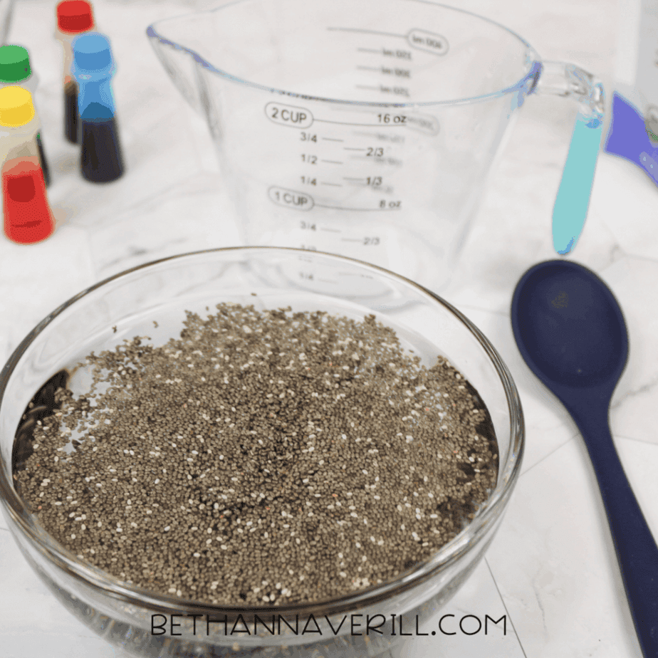 Glass bowl filled with water and dry chia seeds resting on a countertop, with measuring tools and food coloring bottles nearby.