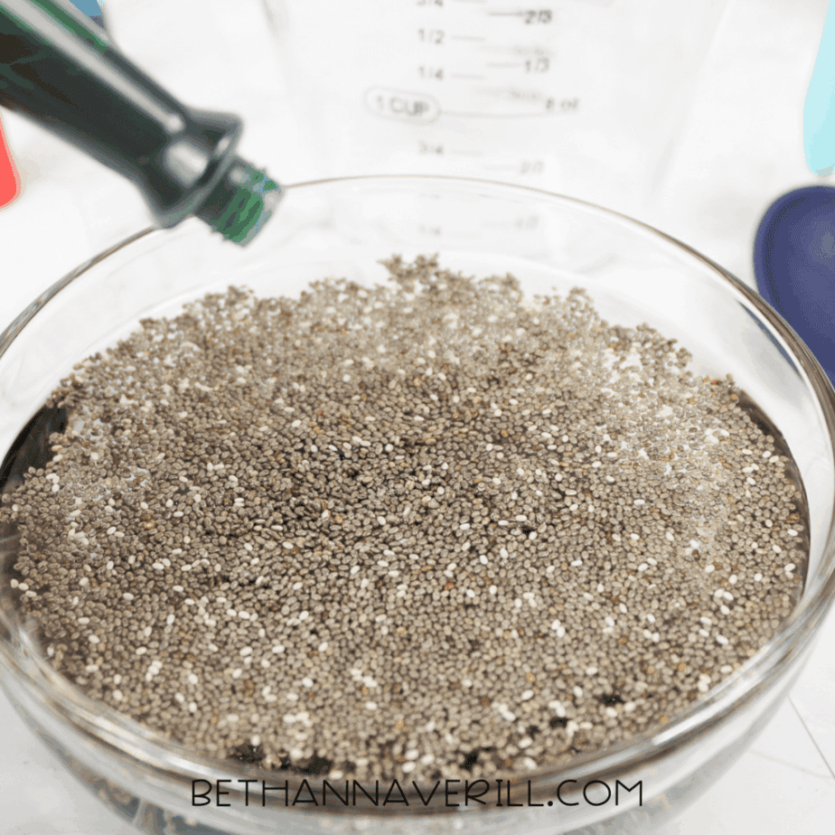 Food coloring bottle pouring liquid into a glass bowl filled with chia seeds and water.