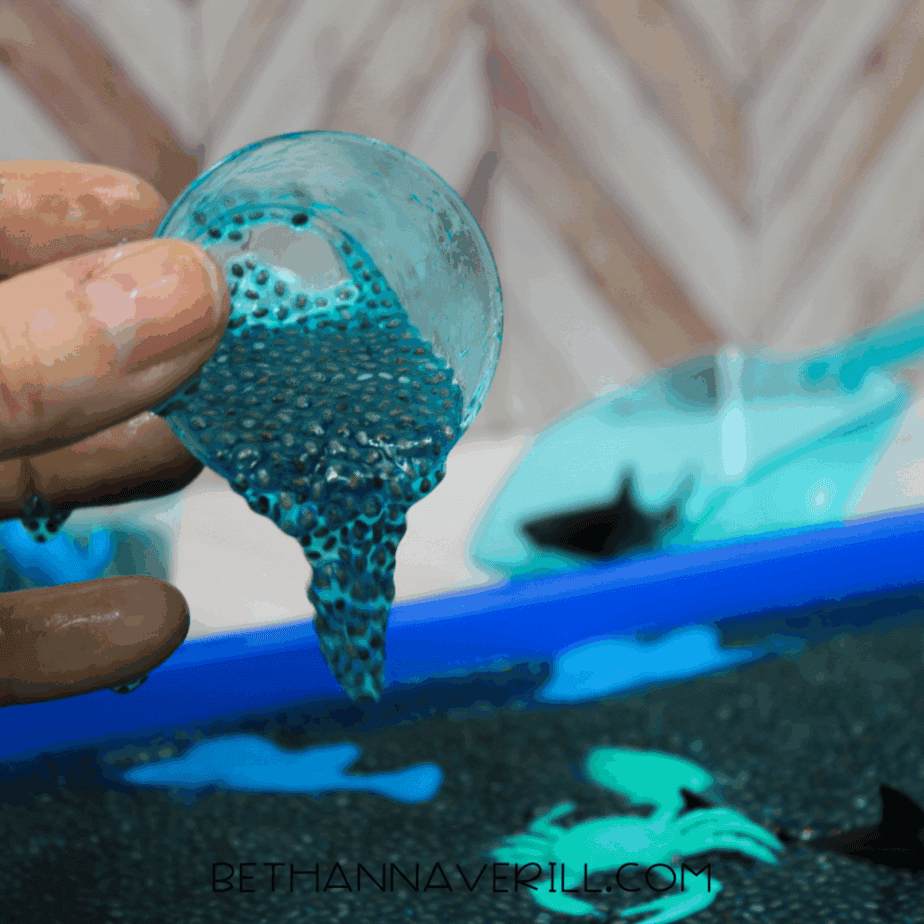 Close-up of blue hydrated chia seeds being poured from a small clear cup into the sensory bin with ocean animal toys visible below.