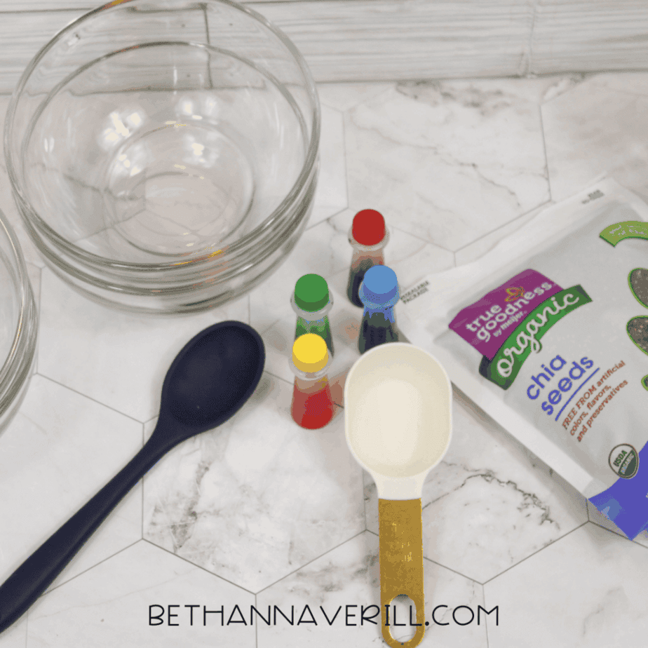 Overhead view of glass bowls, a large spoon, a measuring cup, bottles of liquid food coloring, and a bag of organic chia seeds arranged on a white surface.