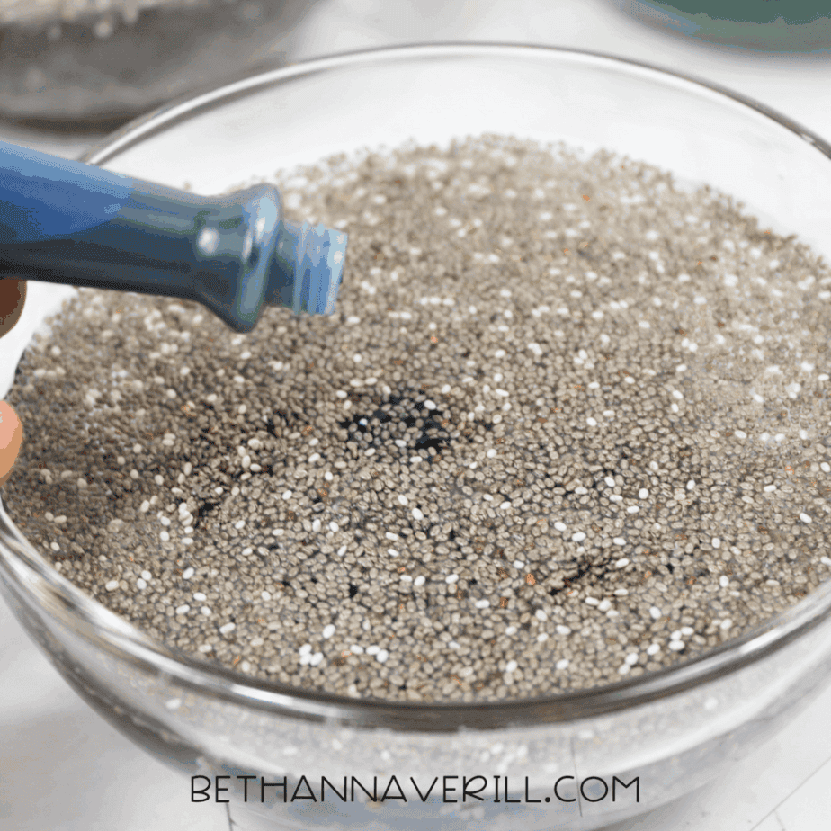 Close-up of blue liquid food coloring being dropped onto hydrated chia seeds in a glass bowl.