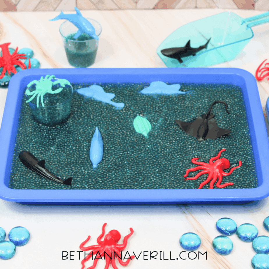 Blue plastic sensory bin filled with blue chia seeds and plastic ocean animals, including sharks, rays, and octopus figures.