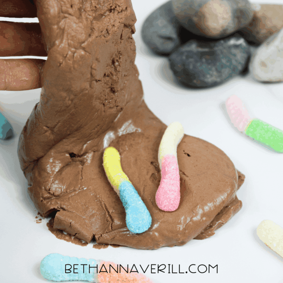 Brown slime being stretched by a hand, with colorful gummy worms pressed into the surface and rocks in the background.