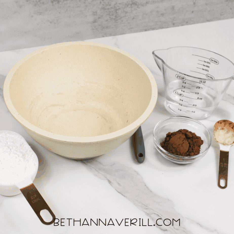 Large mixing bowl on a marble surface next to a measuring cup with water, a small bowl of cocoa powder, and measuring cups containing white powder and cocoa powder.