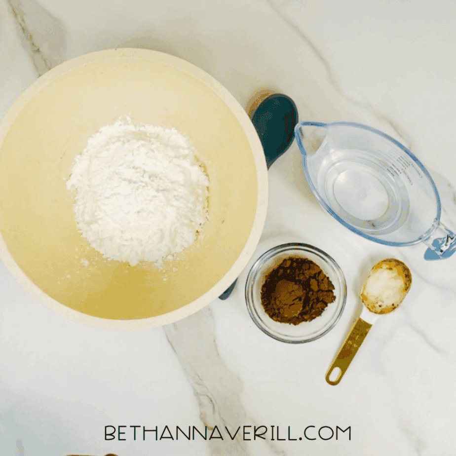 White powder added to a large mixing bowl, with a measuring cup of water, a small bowl of cocoa powder, and measuring cups nearby on a marble surface.