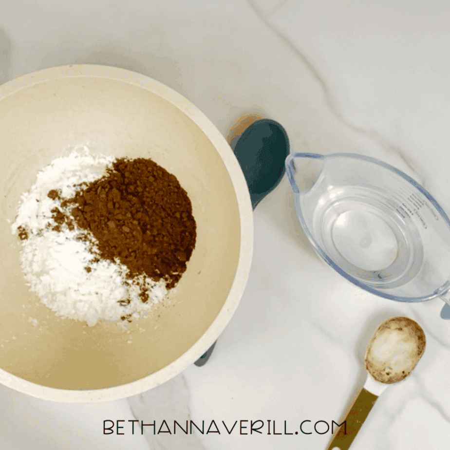 Cocoa powder added on top of white powder inside a large mixing bowl, with a measuring cup of water and a measuring spoon beside the bowl.