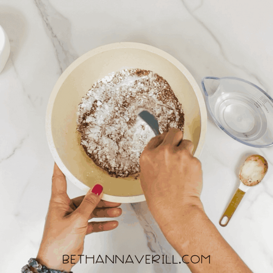 Hands stirring white powder and cocoa powder together inside a large mixing bowl using a spoon on a marble surface.