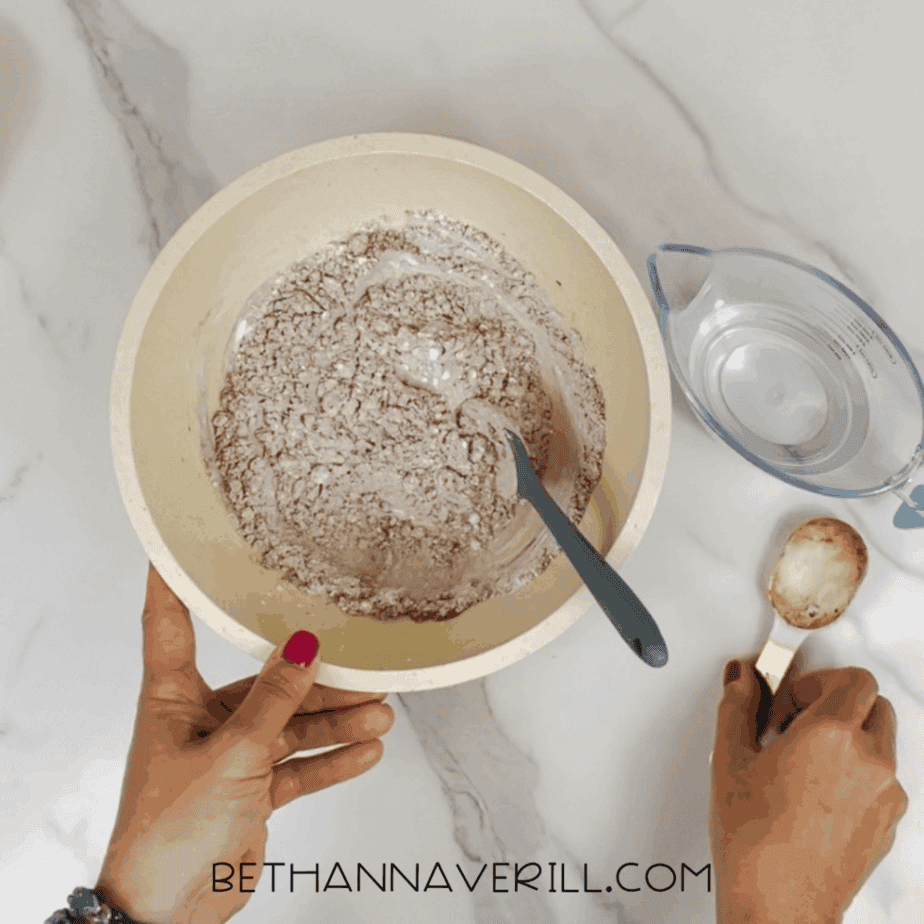 Mixed dry ingredients inside a large bowl, with a spoon resting in the mixture and a measuring cup of water and measuring spoon nearby.