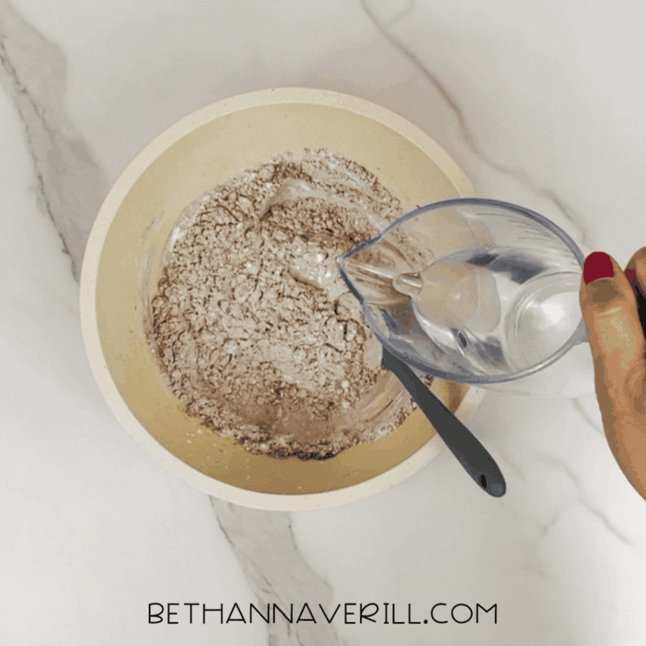 Water being poured from a measuring cup into a bowl containing mixed dry ingredients, with a spoon inside the bowl.
