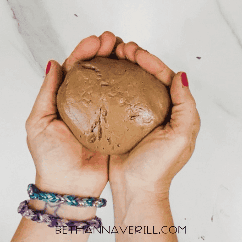 Two hands holding a smooth brown dough-like mixture formed into a ball over a marble surface.