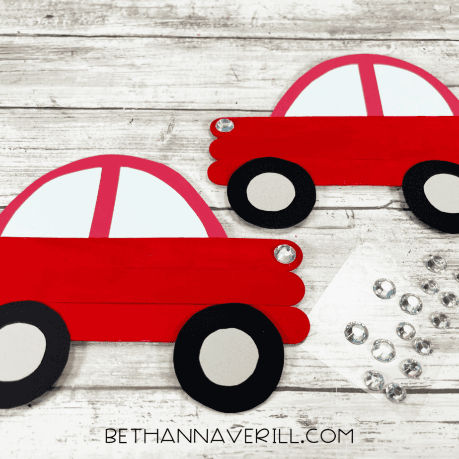Two completed popsicle stick car crafts with red bodies, white windows, black wheels, and rhinestone details displayed on a white wood background.