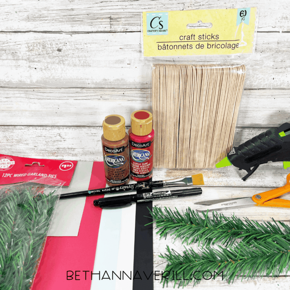 Dollar store craft supplies for a Christmas car popsicle stick craft including jumbo craft sticks, acrylic paint bottles, paintbrush, black marker, scissors, cardstock, wired garland, and a hot glue gun on a white wood background.