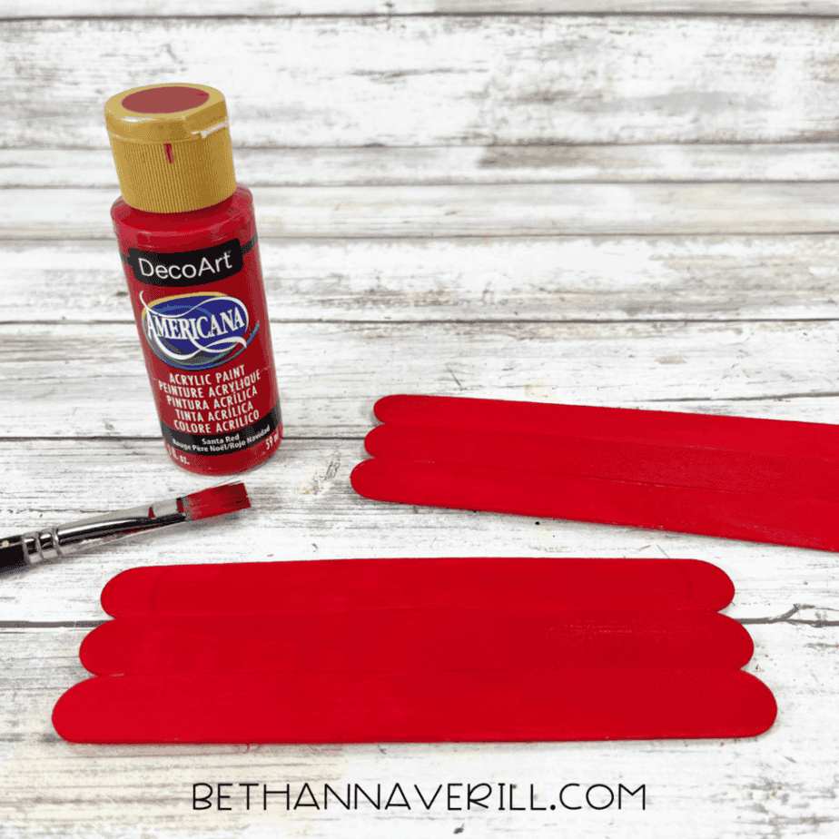 Popsicle sticks painted red laid flat on a white wood background with a red acrylic paint bottle and paintbrush.