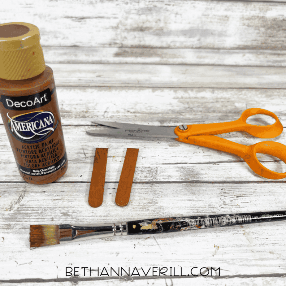 Brown painted craft stick pieces shown with a paintbrush, scissors, and acrylic paint on a white wood background.