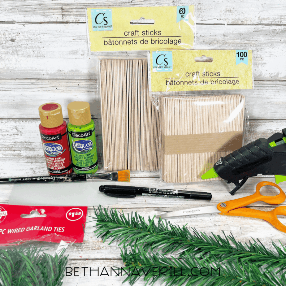 Dollar store supplies for a Christmas door popsicle stick craft including craft sticks, acrylic paint, scissors, glue gun, faux greenery, and ribbon