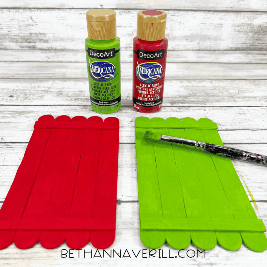 Painted red and green popsicle stick doors drying with acrylic paint and a paintbrush nearby