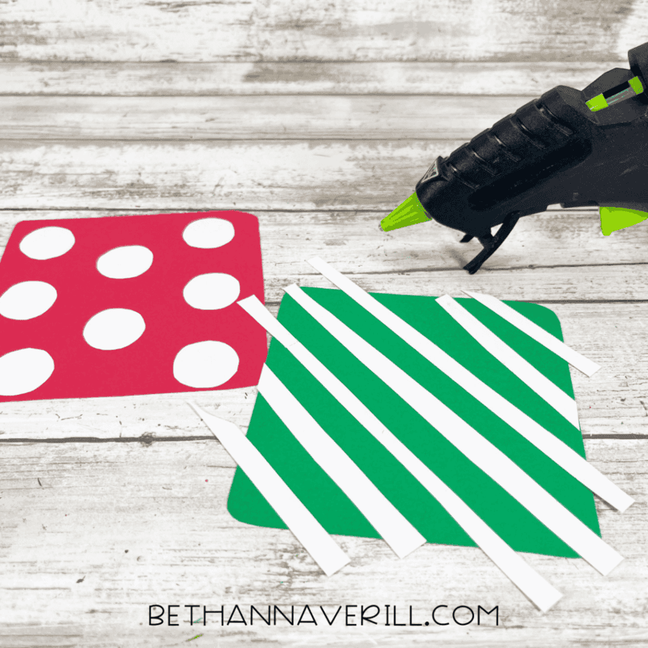 Red polka-dot cardstock square and green striped cardstock square on a white wood surface with white paper strips placed on top and a black glue gun to the right.
