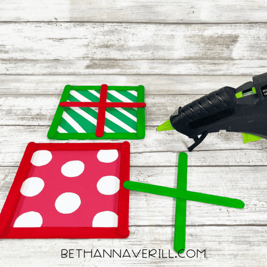 Painted craft stick frames in red and green surrounding patterned cardstock squares, with additional painted craft sticks forming cross shapes and a glue gun on a white wood surface.
