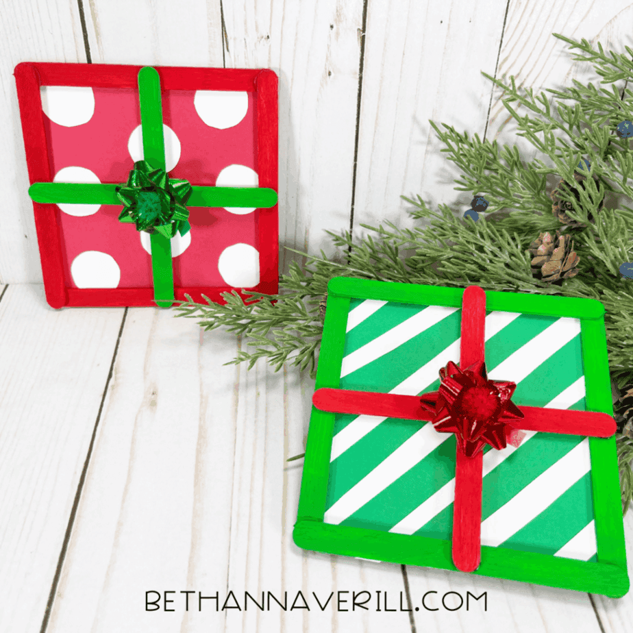 Two finished popsicle stick gift decorations displayed upright on a white wood surface with faux greenery, pinecones, and berries arranged around them.