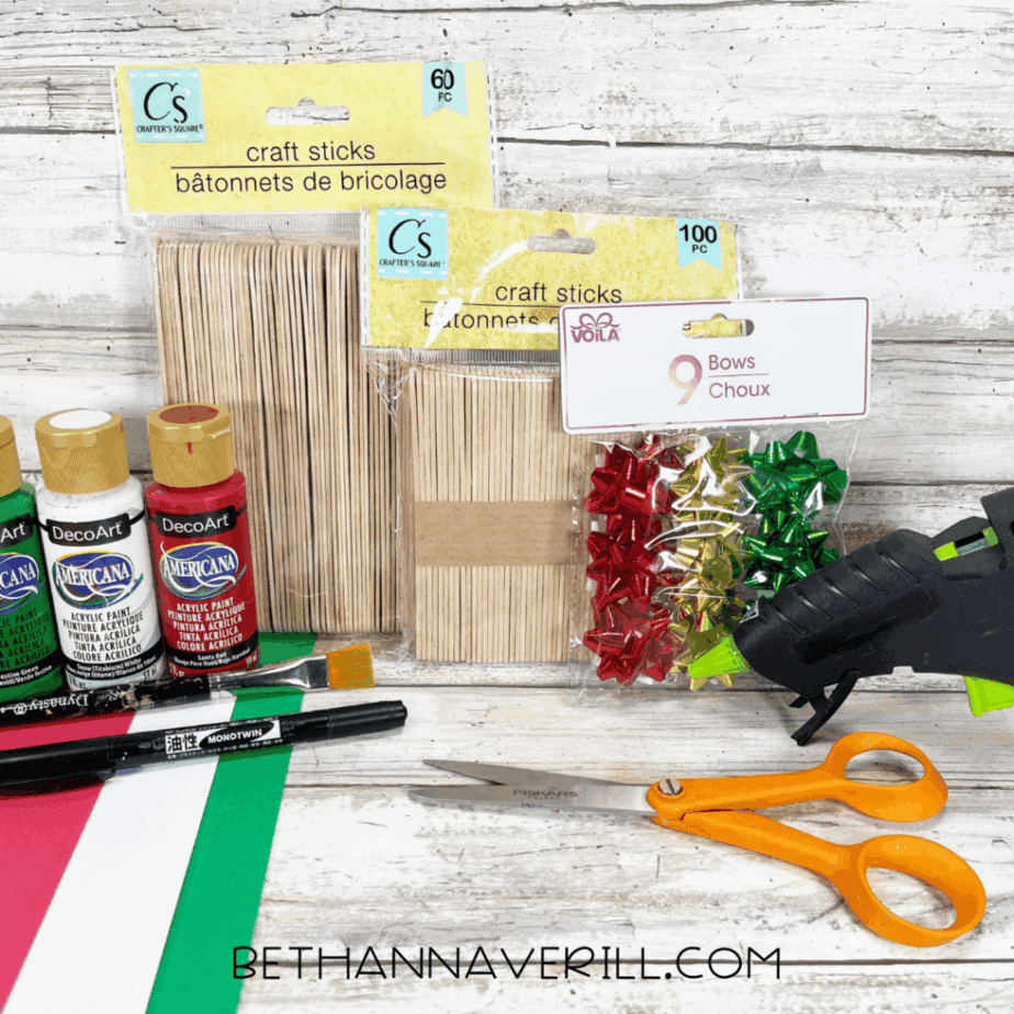 Craft supplies for a Christmas popsicle stick gift craft, including craft sticks, acrylic paint bottles, small gift bows, scissors, a glue gun, paintbrushes, marker, cardstock, and ribbon laid out on a white wood surface.
