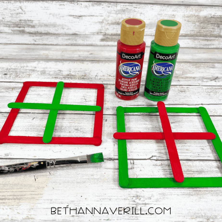 Painted craft stick frames in red and green arranged in square shapes, with red and green acrylic paint bottles and a paintbrush on a white wood background.