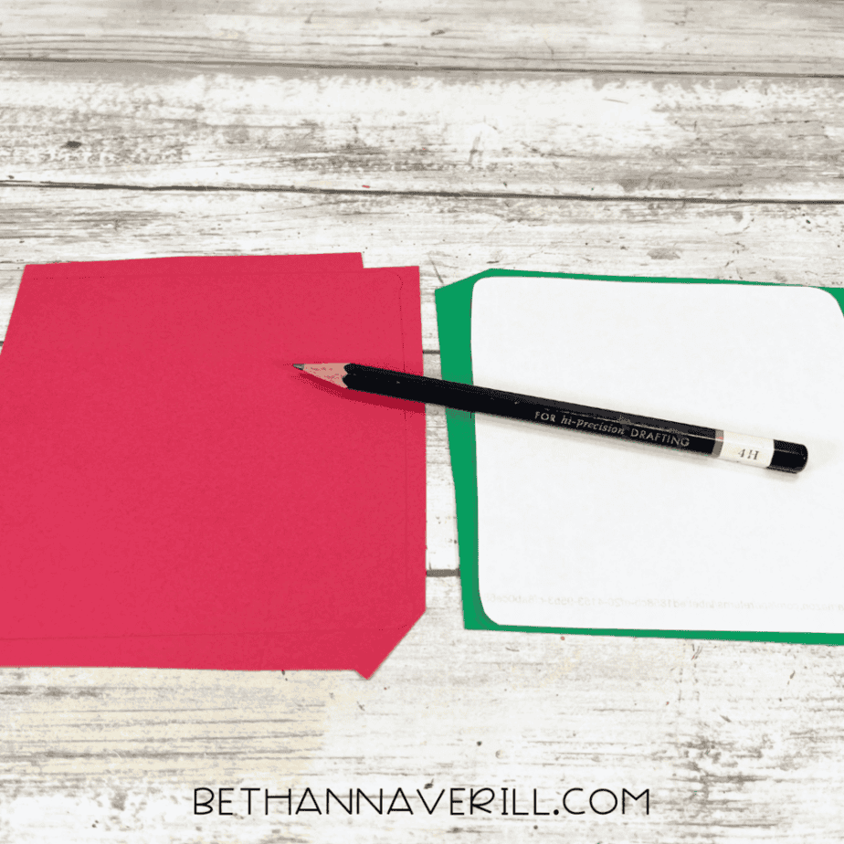 Red and green cardstock with rounded corners being traced using a pencil on a white wood surface.
