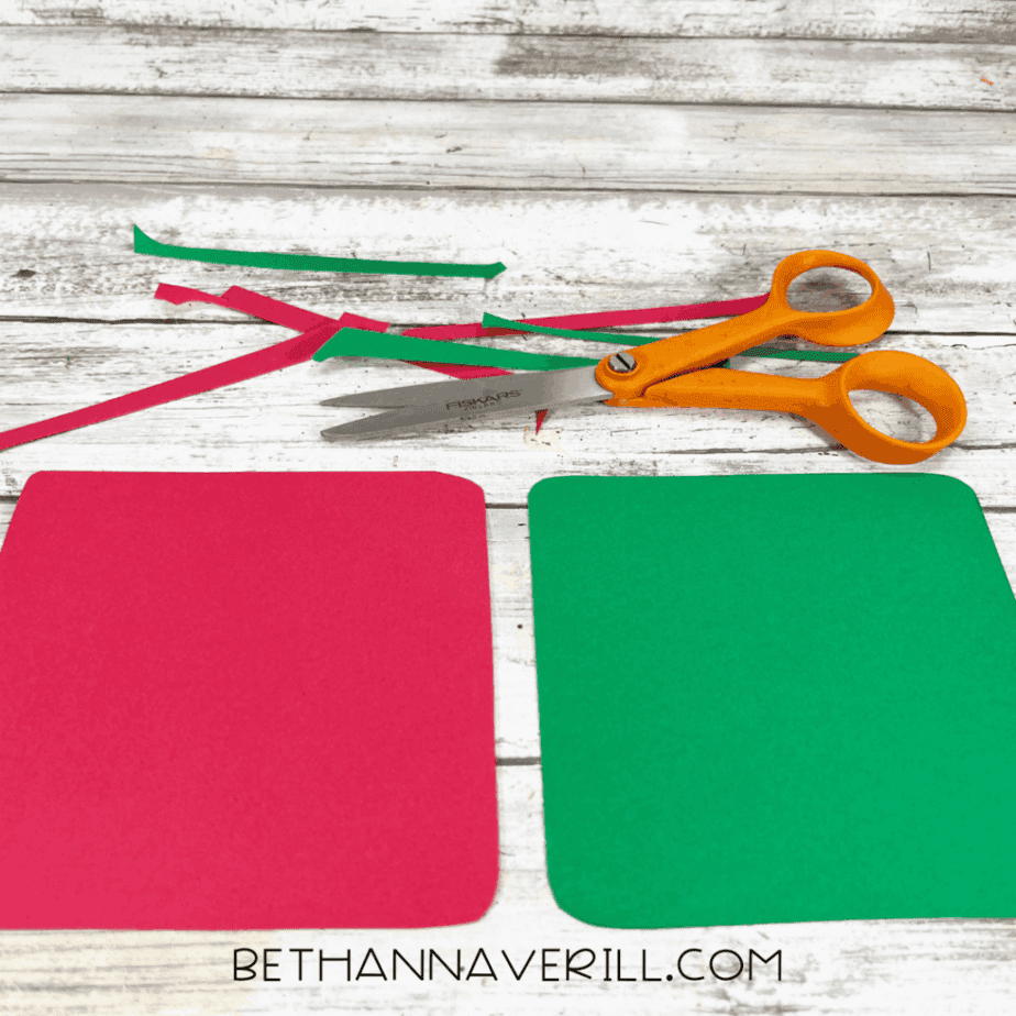 Red and green cardstock squares with rounded corners next to thin strips of cardstock and scissors on a white wood background.