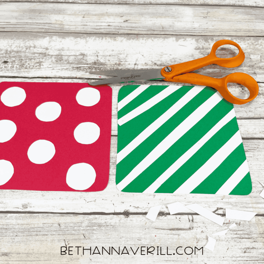 Two square cardstock pieces on a white wood surface, one red with white circles and one green with diagonal white stripes, with orange-handled scissors above them.