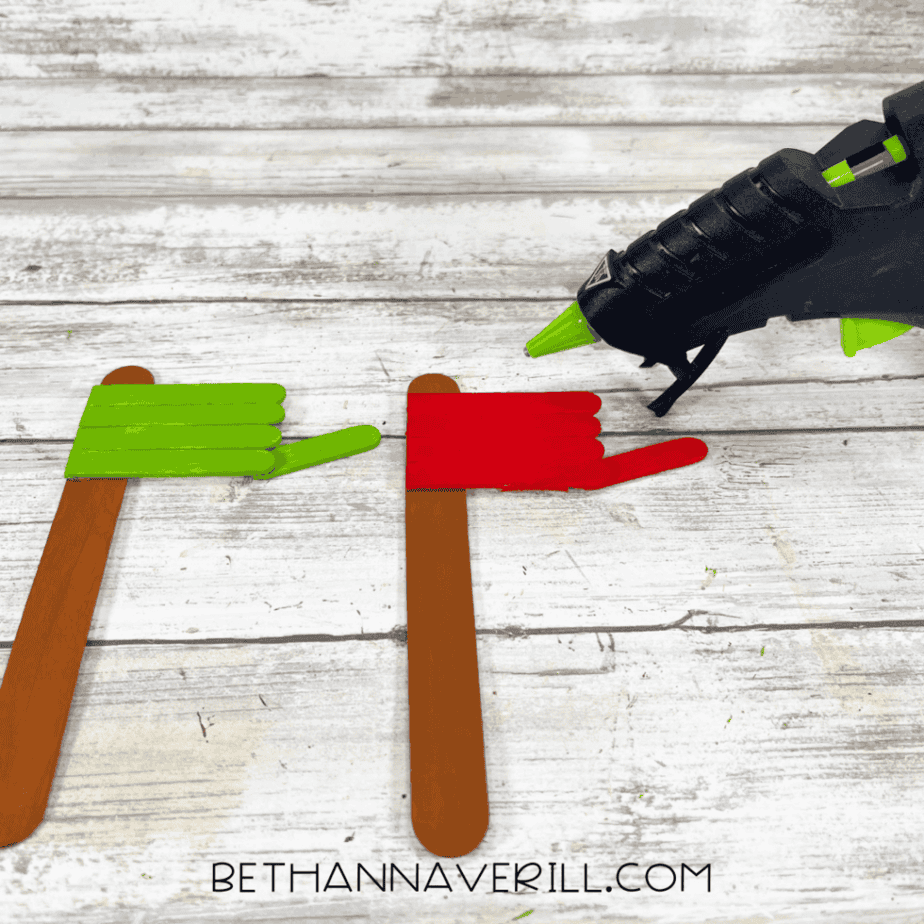 Two painted popsicle stick mailbox bases, one green and one red, attached to brown craft stick posts, with a glue gun positioned to the right on a white wood background.