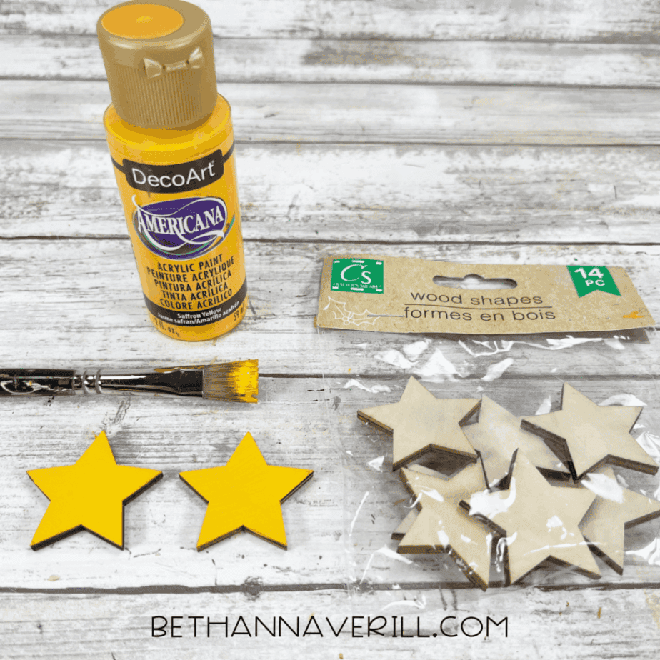Craft supplies for a Christmas window popsicle stick craft including yellow acrylic paint, a paintbrush, wooden star shapes, and painted yellow stars on a white wood surface.
