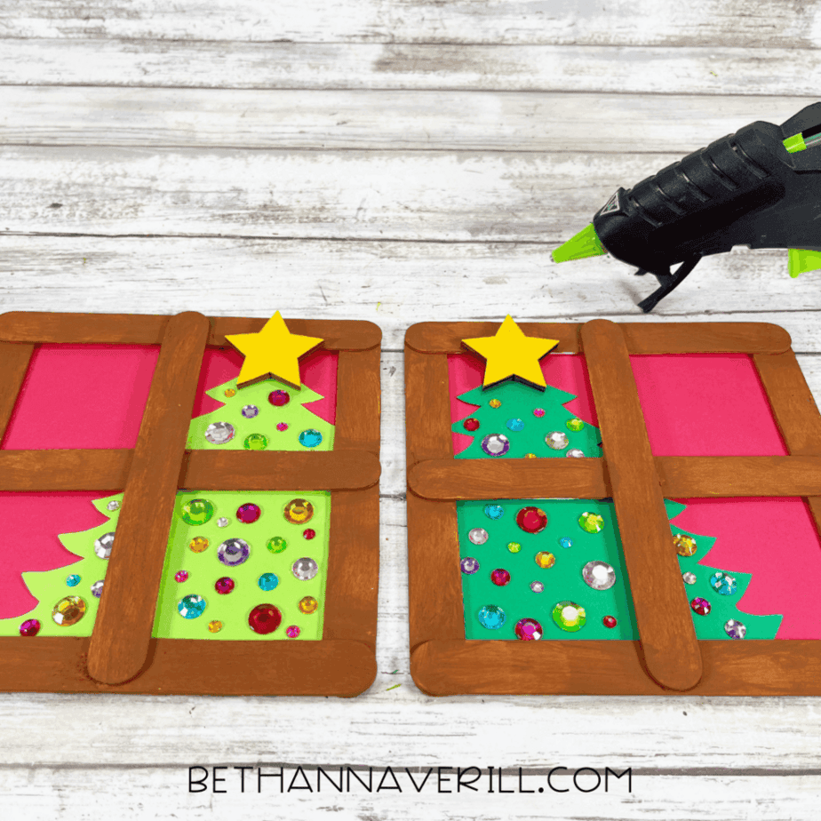 Two popsicle stick window frames painted brown placed over pink cardstock backgrounds with green Christmas trees decorated with gem stickers, with a glue gun to the right.