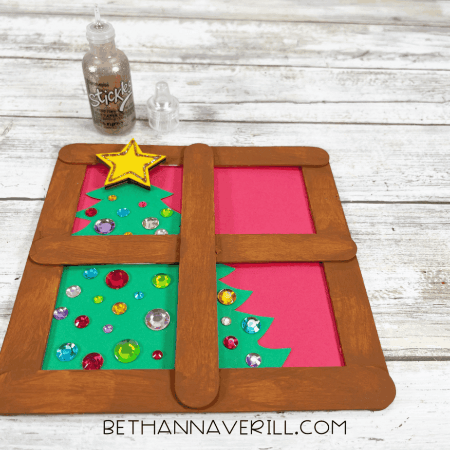 One completed Christmas window popsicle stick craft showing a brown window frame, a green cardstock Christmas tree with colorful gem stickers, and a yellow star on a pink background.