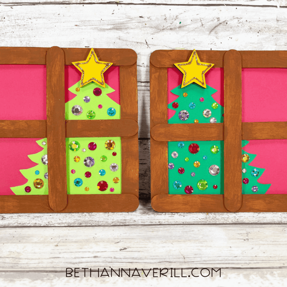 Two completed Christmas window popsicle stick crafts displayed side by side, each with brown frames, green cardstock trees decorated with gem stickers, and yellow stars.