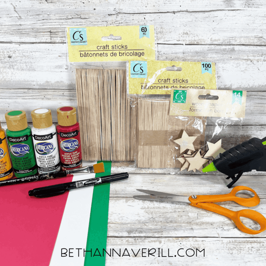 Craft supplies for a Christmas window popsicle stick craft including craft sticks, acrylic paint bottles, cardstock, scissors, a paintbrush, a black marker, wooden stars, and a glue gun on a white wood surface.