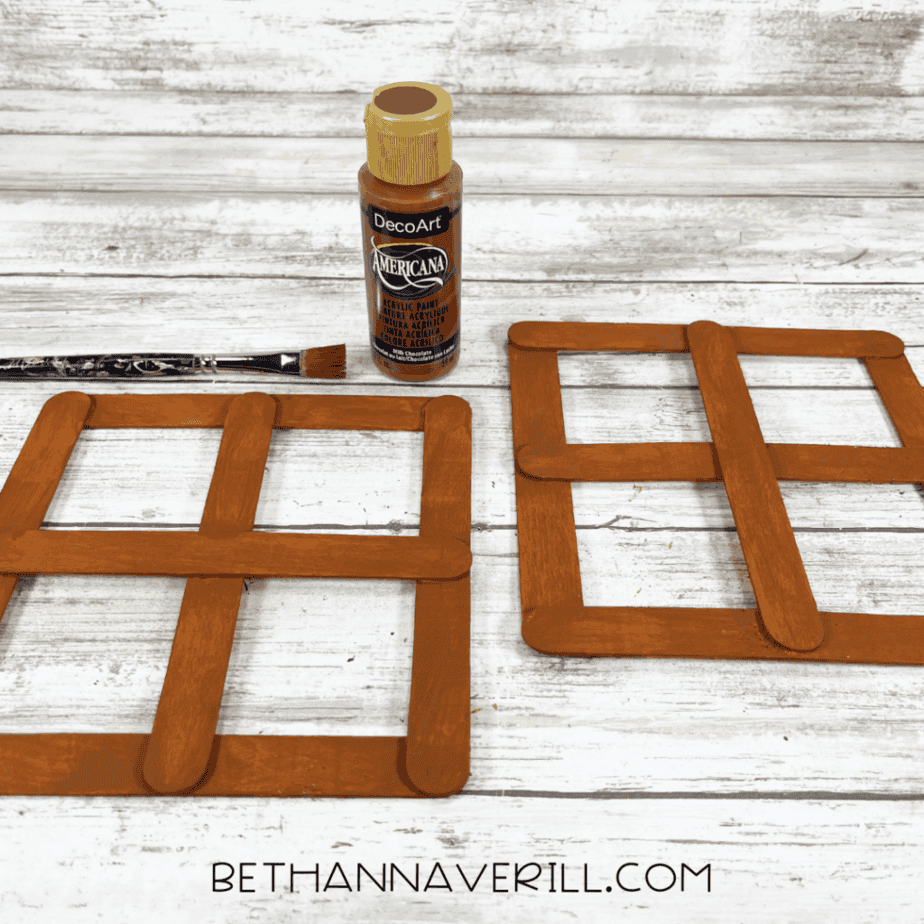 Two popsicle stick window frames painted brown, with a bottle of brown acrylic paint and a paintbrush above them on a white wood background.